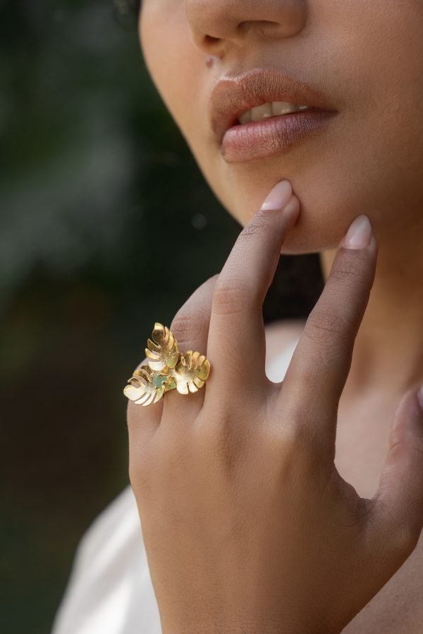 A close-up of a woman’s hand touching her chin, wearing a large gold leaf-shaped ring on her finger.