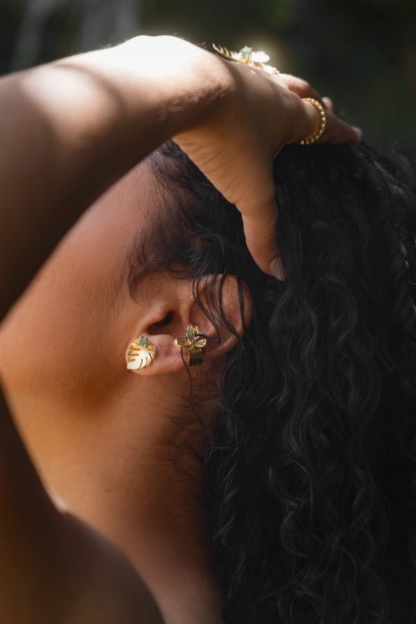 Close-up of a person with curly hair, wearing gold earrings shaped like a leaf and a flower, and gold rings, with one hand resting on their head.