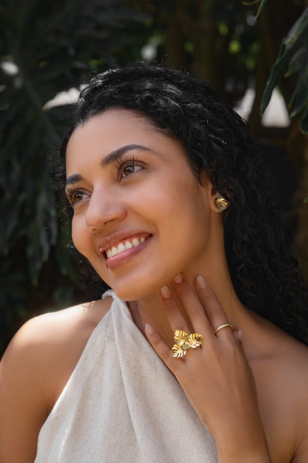 A woman with curly hair wearing a sleeveless top smiles, showing gold jewelry including a ring, earrings, and a small gold band. Green foliage is in the background.