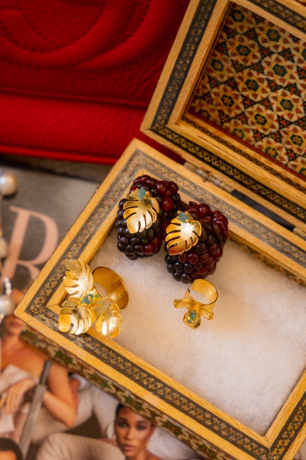 A decorative jewelry box displays a pair of gold leaf earrings with green stones and matching gold rings. The background includes a red item and a partially visible magazine.